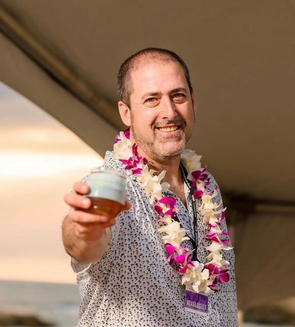 Man holding a cup outdoors wearing a purple and white single-strand orchid lei, with a blurred natural background.