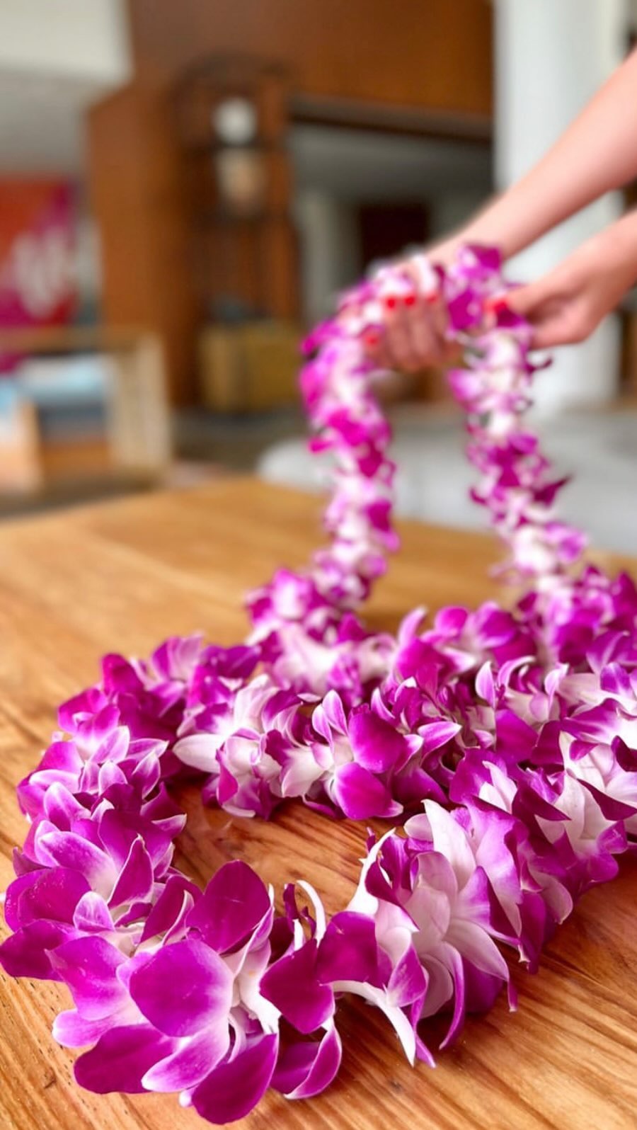 Two single-strand purple orchid leis resting on a wooden table, with hands holding one lei blurred in the background.