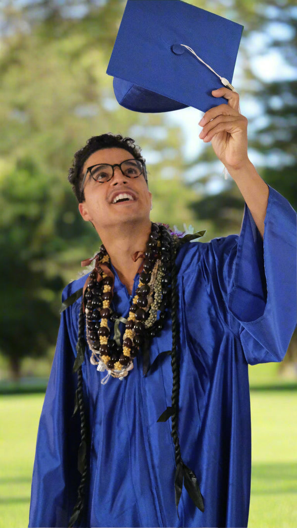 Graduate in blue cap and gown celebrating outdoors with mongo shell, kukui, and single ti leaf leis.
