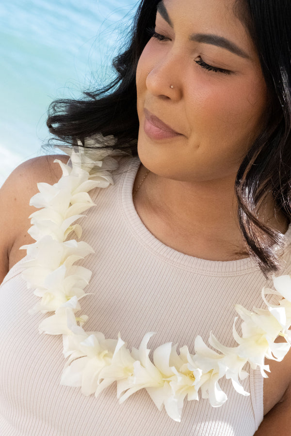 A girl wearing a beige tank top and a pure white orchid lei, standing with the ocean in the background.