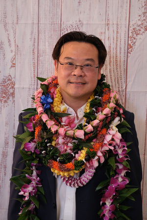 The owner of the orchid lei company adorned with many leis, standing in front of a wooden background.