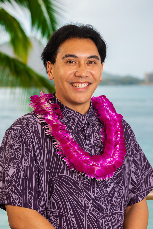 A man wearing a two-tone purple patterned shirt and a fluffy purple orchid lei in front of Diamond Head, with a palm tree and the ocean in the background.