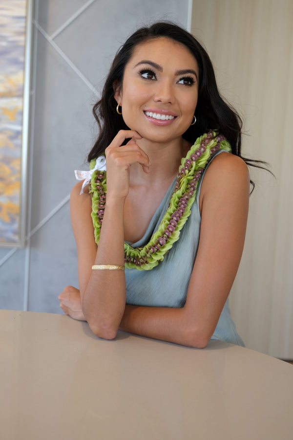 Woman wearing a teal dress and an all jade orchid lei with dark purple centers, accented with a white bow, resting her arm on a table.
