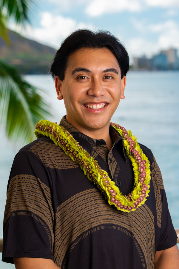 Man wearing a black and brown patterned shirt and an all jade orchid lei with dark purple centers, with Diamond Head, a palm tree, and the ocean in the background.