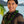 Man wearing a ti leaf kukui nut lei with folded green ti leaves, shiny black kukui nuts, and white mongo shells, set against a tropical background.