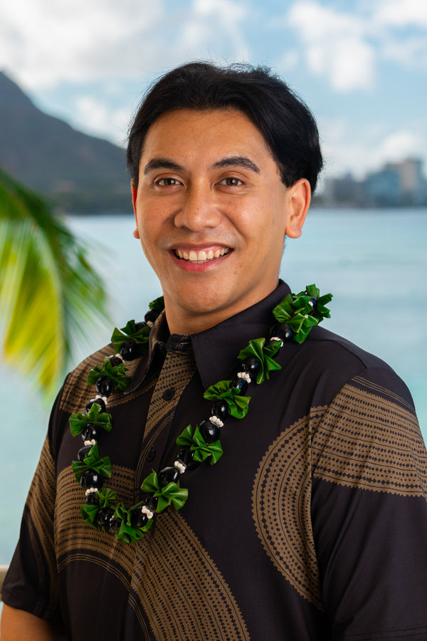 Man wearing a ti leaf kukui nut lei with folded green ti leaves, shiny black kukui nuts, and white mongo shells, set against a tropical background.