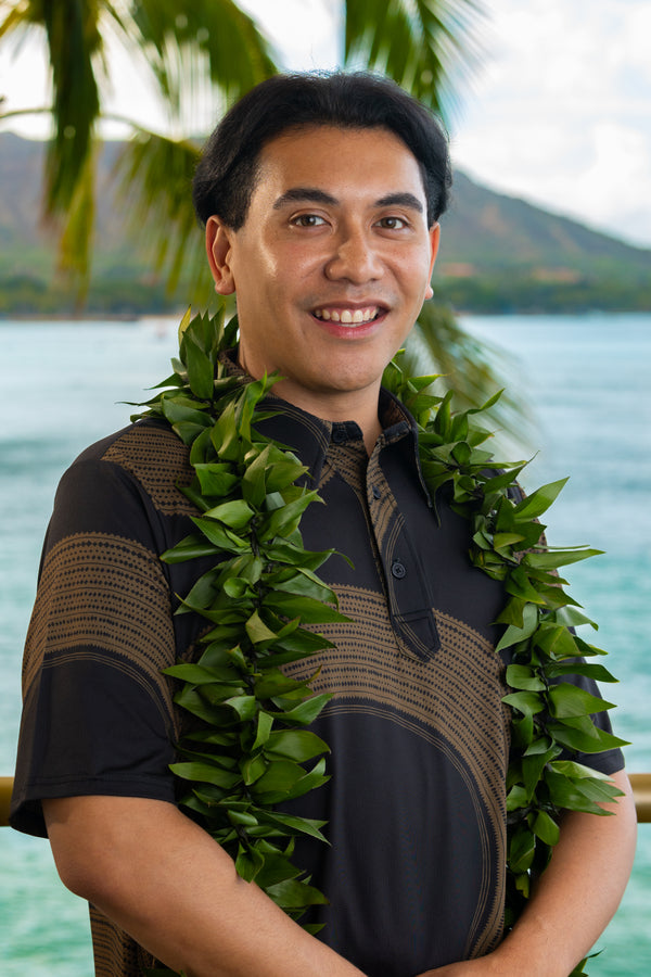 Man wearing a fresh and lush green ti leaf maile lei with Diamond Head, a palm tree, and the ocean in the background.
