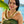Girl in her 20s wearing a dark green dress with black flowers and a green and white flat deluxe orchid lei, with Diamond Head, a palm tree, and the ocean in the background.