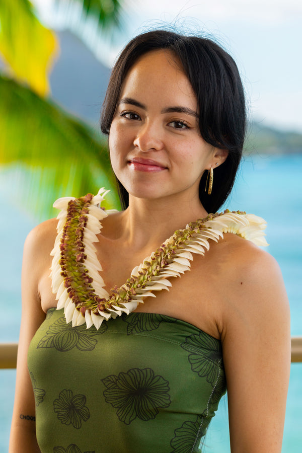 Girl in her 20s wearing a dark green dress with black flowers and a green and white flat deluxe orchid lei, with Diamond Head, a palm tree, and the ocean in the background.