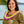 A woman wearing a green leaf pattern shirt and a flat orchid lei featuring purple, white, and green petals with Diamond Head, a palm tree, and the ocean in the background.